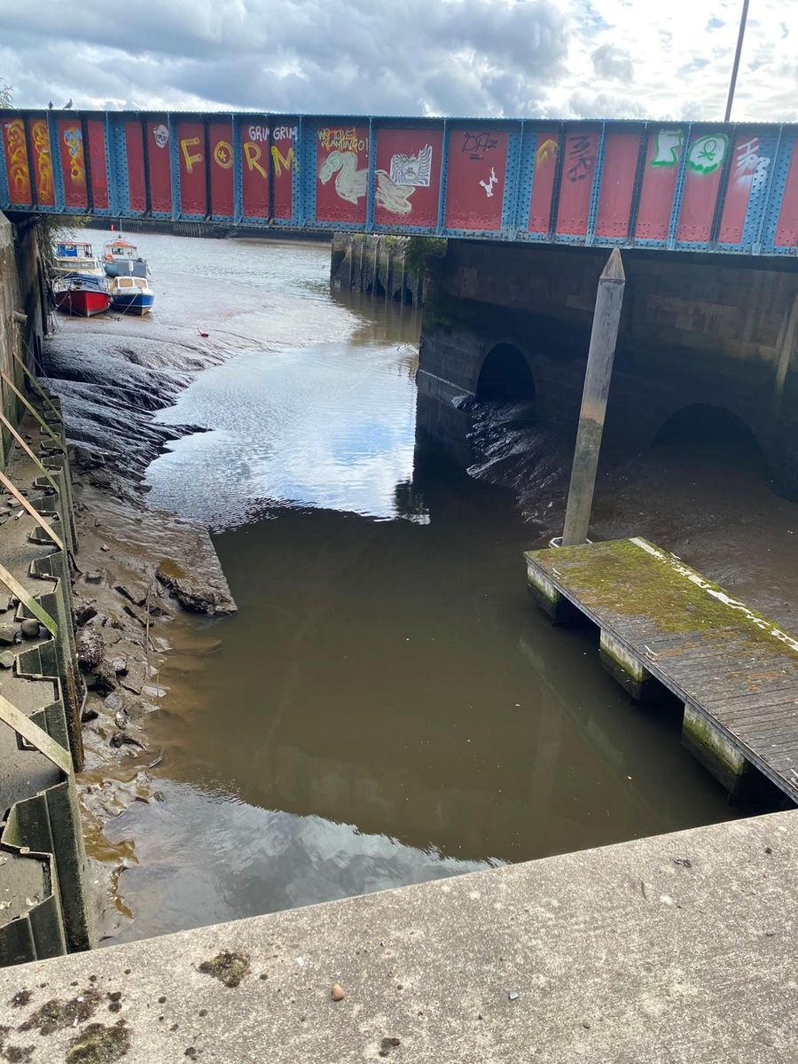 Low tide at the mouth of the Ouseburn, a tributary of the Tyne in Newcastle, showing exposed mudbanks beneath a railway bridge with small boats moored nearby.